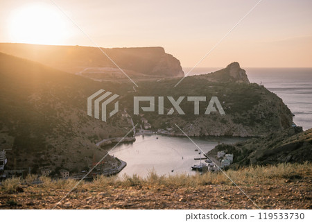 A beautiful sunset over a mountain with a small town in the distance. The sun is setting behind the mountains, casting a warm glow over the landscape. The water in the valley is calm and peaceful. A beautiful sunset over a mountain with a small town in the distance. The sun is setting behind the mountains, casting a warm glow over the landscape. The water in the valley is calm and peaceful. 119533730