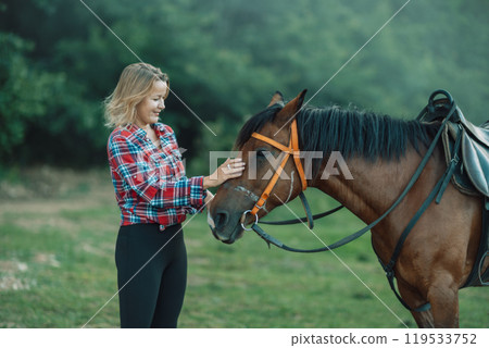 Happy blonde with horse in forest. Woman and a horse walking through the field during the day. Dressed in a plaid shirt and black leggings. 119533752
