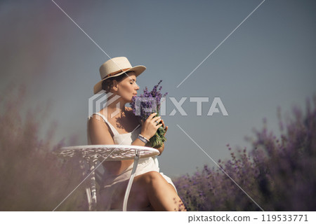 woman sitting in a field of lavender and wearing a straw hat. She is smiling and holding a bouquet of flowers. Scene is peaceful and serene, as the woman is surrounded by the beauty of nature 119533771