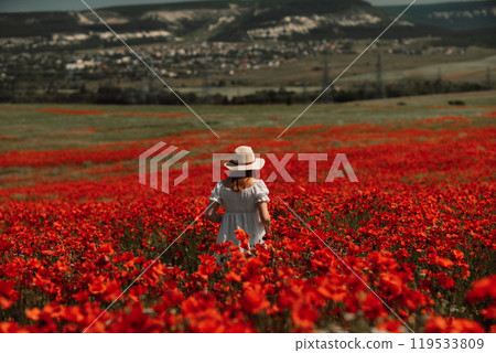 Field poppies woman. Happy woman in a white dress and hat stand with her back a blooming field of poppy. Field of blooming poppies. 119533809