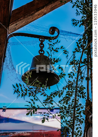 Weathered vintage bell dangles from sturdy wooden structure in lush green leaves 119533946