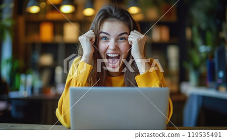Successful Young Business Woman In Her 20s, Happily Working On Laptop At Wooden Office Desk In Casual Yellow Shirt. Celebrating Career Achievement. 119535491