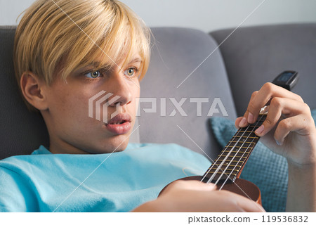 Teen boy focuses on playing ukulele guitar, capture moment of musical exploration. Teen boy focuses on playing ukulele guitar, capture moment of musical exploration. 119536832