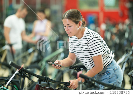 Young girl using smartphone for examining quality seat and choosing bike in bicycle sports store 119538735