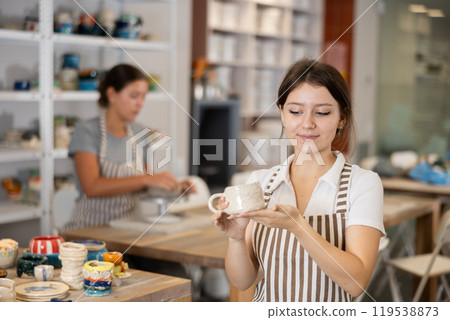 Girl stands in pottery workshop and shows created work, finished decorative vase - jug. Girl stands in pottery workshop and shows created work, finished decorative vase - jug. 119538873