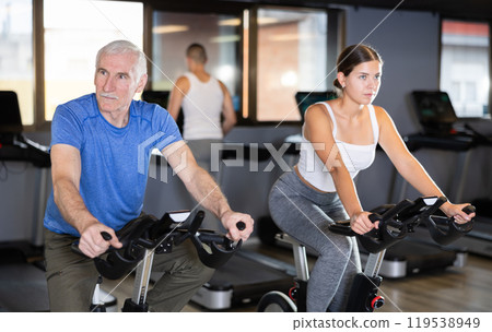 Elderly man, along with other people, is engaged on an bike in gym Elderly man, along with other people, is engaged on an bike in gym 119538949
