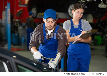 Female car service manager makes entries in a journal while monitoring the work of an auto mechanic repairing car 119538957