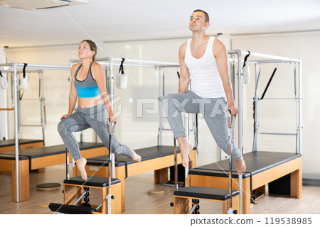 Young man and woman performing Pilates exercises on Wunda chairs Young man and woman performing Pilates exercises on Wunda chairs 119538985