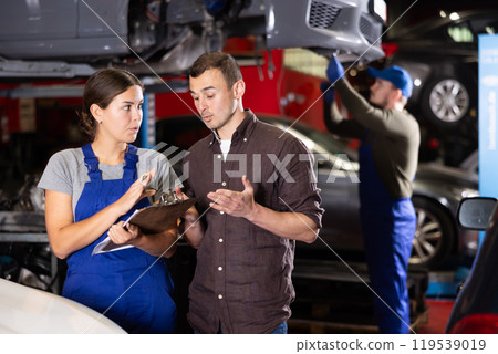 Female worker of auto service speaking to male client concerning her notes on paper Female worker of auto service speaking to male client concerning her notes on paper 119539019