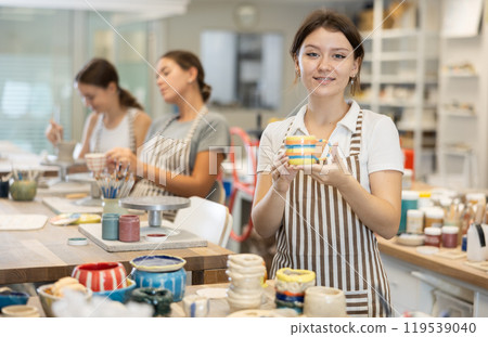 Girl stands in pottery workshop and shows created work, finished decorative vase - jug. Girl stands in pottery workshop and shows created work, finished decorative vase - jug. 119539040