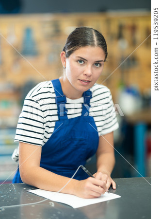 Polite young girl salesperson dressed in uniform taking order and writing form in sports store Polite young girl salesperson dressed in uniform taking order and writing form in sports store 119539205
