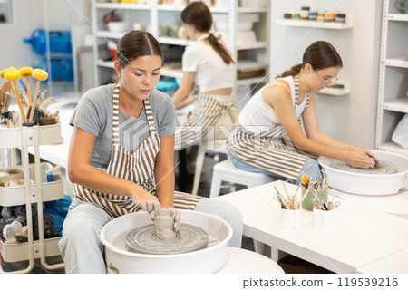 Three young women sits near potter wheel and makes clay crafts Three young women sits near potter wheel and makes clay crafts 119539216