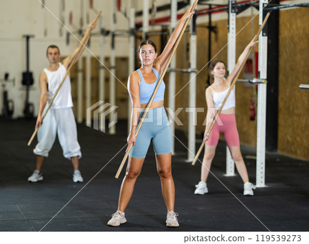 Slender girl, together with other athletes, does a warm-up using a long stick in gym Slender girl, together with other athletes, does a warm-up using a long stick in gym 119539273