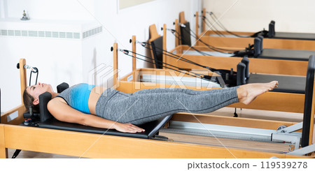 Girl works out on in gym on Pilates simulator. Sports club client on reformer with cable and rope performs exercise to stretch muscles of arms in gym Girl works out on in gym on Pilates simulator. Sports club client on reformer with cable and rope performs exercise to stretch muscles of arms in gym 119539278