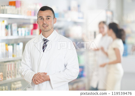 Positive male pharmacist in medical uniform posing while working in pharmacy Positive male pharmacist in medical uniform posing while working in pharmacy 119539476