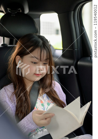 A young, adorable Asian girl is focused on reading a book while on her way to school. 119540012