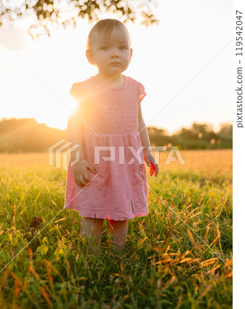 Baby girl staying in grass with sunset tones. Cute child in dress outdoor 119542047