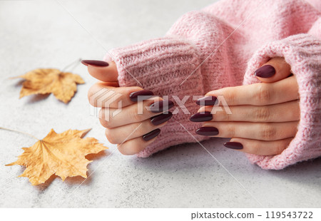 Stylish manicure with dark nails against a cozy pink sweater and autumn leaves on a textured surface Stylish manicure with dark nails against a cozy pink sweater and autumn leaves on a textured surface 119543722