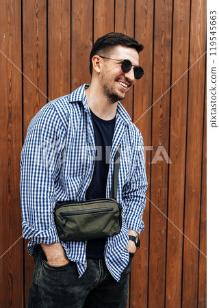A man in a blue checkered shirt smiles while standing outside a modern building on a sunny day A man in a blue checkered shirt smiles while standing outside a modern building on a sunny day 119545663