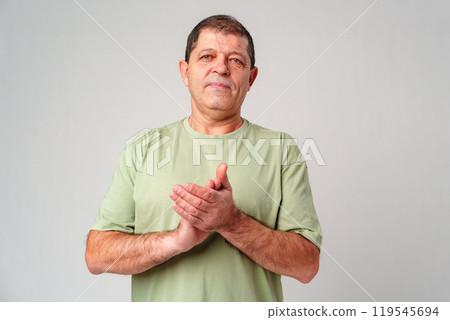 Elderly man claps his hands while standing against a neutral background in a casual light green shirt 119545694