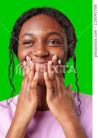 A young woman expresses joy while smiling and touching her face against a bright green background A young woman expresses joy while smiling and touching her face against a bright green background 119545709