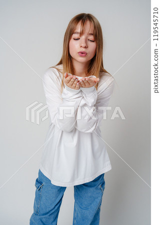 Young woman in white long-sleeve shirt blowing a kiss against a plain background 119545710