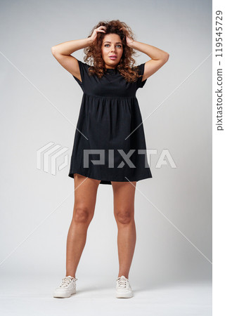 Smiling woman with curly hair poses confidently against a plain background while holding her hands behind her head Smiling woman with curly hair poses confidently against a plain background while holding her hands behind her head 119545729