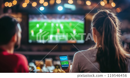 A group of friends watch a live soccer match on a large screen, with one person checking a sports app on their phone. 119546456