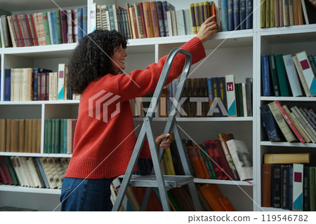 Curly-haired young girl in red jacket on the ladder in the library 119546782