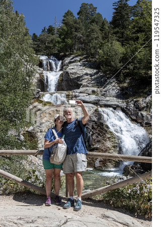 Backpacker middle old tourist couple taking photos of themselves in front of a waterfall in Spanish mountain. 119547325