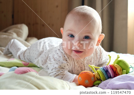Smiling baby girl learning to crawl and playing with colorful toys in white sunny bedroom. 119547470