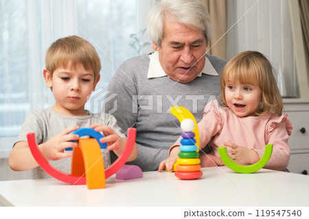 Grandfather playing toys with grandson and young granddaughter indoor. Old man grandpa studying with his grandchildren at weekend. Senior male having fun with kids. Happy multigeneration family video 119547540