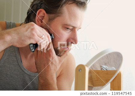 Reflection of a young bearded man shaving his beard holding electric shaver in bathroom. Handsome man sitting in front of the mirror. 30s male cutting trimming mustache using trimmer. Morning routine Reflection of a young bearded man shaving his beard holding electric shaver in bathroom. Handsome man sitting in front of the mirror. 30s male cutting trimming mustache using trimmer. Morning routine 119547545