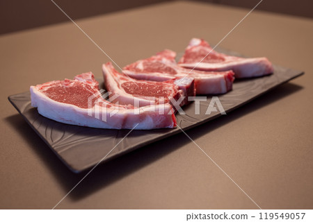 Four raw lamb chops on black plate display pink meat with white fat showing freshness and quality Four raw lamb chops on black plate display pink meat with white fat showing freshness and quality 119549057