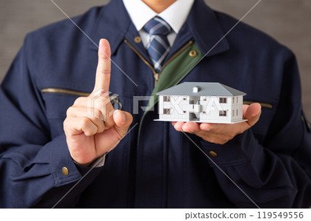 A middle-aged male businessman wearing work clothes holding a model of an apartment building and pointing his index finger 119549556