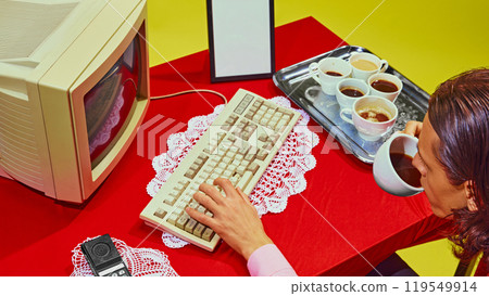 Overhead view of man sitting at red desk drinking coffee while typing on old PC keyboard, tray of coffee cups next to him. 119549914