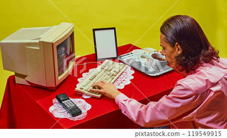 Man sitting at red desk, typing on vintage keyboard while drinking coffee, multiple cups on tray beside him. 119549915