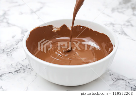 Smooth chocolate ganache being poured from a spoon into a white bowl on a marble countertop in a 119552208