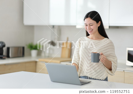 Happy asian woman using laptop drinking coffee in the kitchen Happy asian woman using laptop drinking coffee in the kitchen 119552751