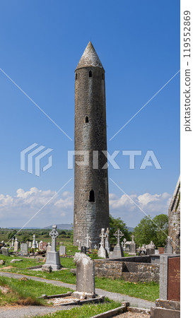 Tall round tower at Kilmacduagh Abbey surrounded by ancient gravestones under bright blue sky 119552869