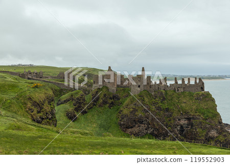 Dunluce Castle ruins atop grassy cliff with ocean view surrounded by green pastures cloudy sky Dunluce Castle ruins atop grassy cliff with ocean view surrounded by green pastures cloudy sky 119552903