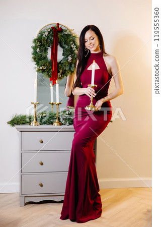 A beautiful brunette woman with makeup in a red dress celebrates the new year in a decorated festive interior of a room with a Christmas tree on a holiday at home 119555360