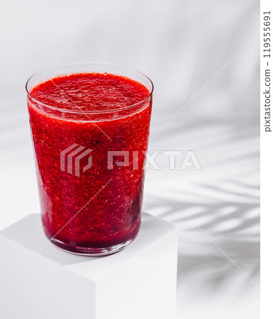 Glass of red berry smoothie sitting on white cube with tropical plant shadow Glass of red berry smoothie sitting on white cube with tropical plant shadow 119555691