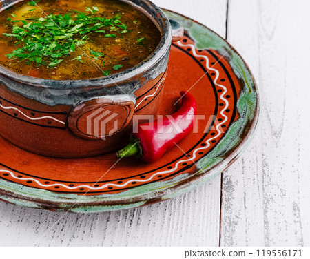 Rustic clay bowl presenting steaming hot soup with fresh herbs and chili pepper 119556171