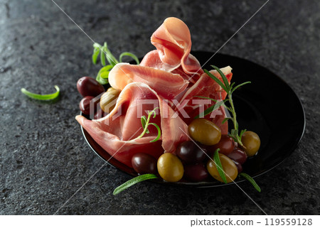 Italian prosciutto crudo with olives and rosemary on a black stone table. Italian prosciutto crudo with olives and rosemary on a black stone table. 119559128