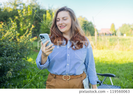Happy Caucasian woman smiling confident using smartphone. Young woman browsing Internet on a phone, texting and communicating outdoors. Travel, fashion, blogging concept. 119560063