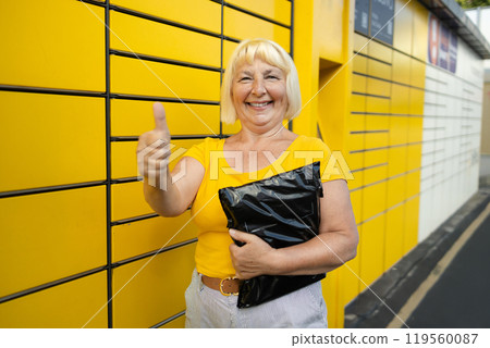 Senior 50s woman showing thumb up getting parcel from cell of automatic post terminal outdoors. Concept of contactless and smart delivery. Idea of modern shipping and logistics. Woman wearing yellow t 119560087