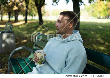 Adult 30s man sits on a bench in an autumn park, drinks coffee or hot tea from a paper glass and looks around. Portrait of a handsome cheerful guy sitting on a bench outdoors using a device browsing 119560099