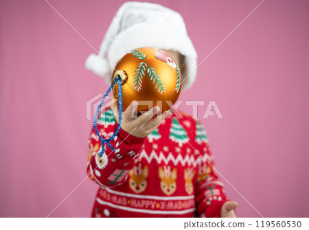 A child in a Santa hat holds a Christmas ornament on a pink background, conveying festive warmth and fun. Christmas card for the holiday. Evokes childish joy and celebration. 119560530