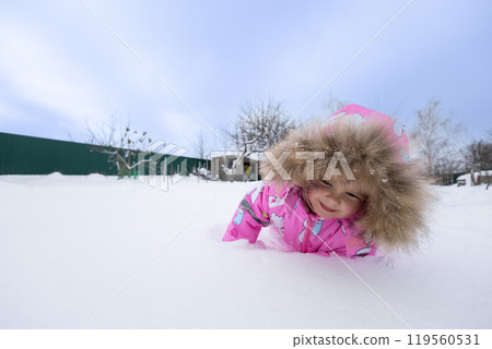 A child explores the snow-covered ground, crawls and plays on a winter day in the backyard. A joyful child plays in the soft snow in winter and enjoys a carefree childhood. Concept child, winter. 119560531